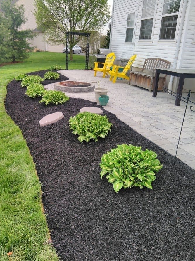 Outdoor Splendor - A landscaped backyard patio with yellow chairs, a fire pit, paver stones, green shrubs, and a mulched garden bed next to a white house.