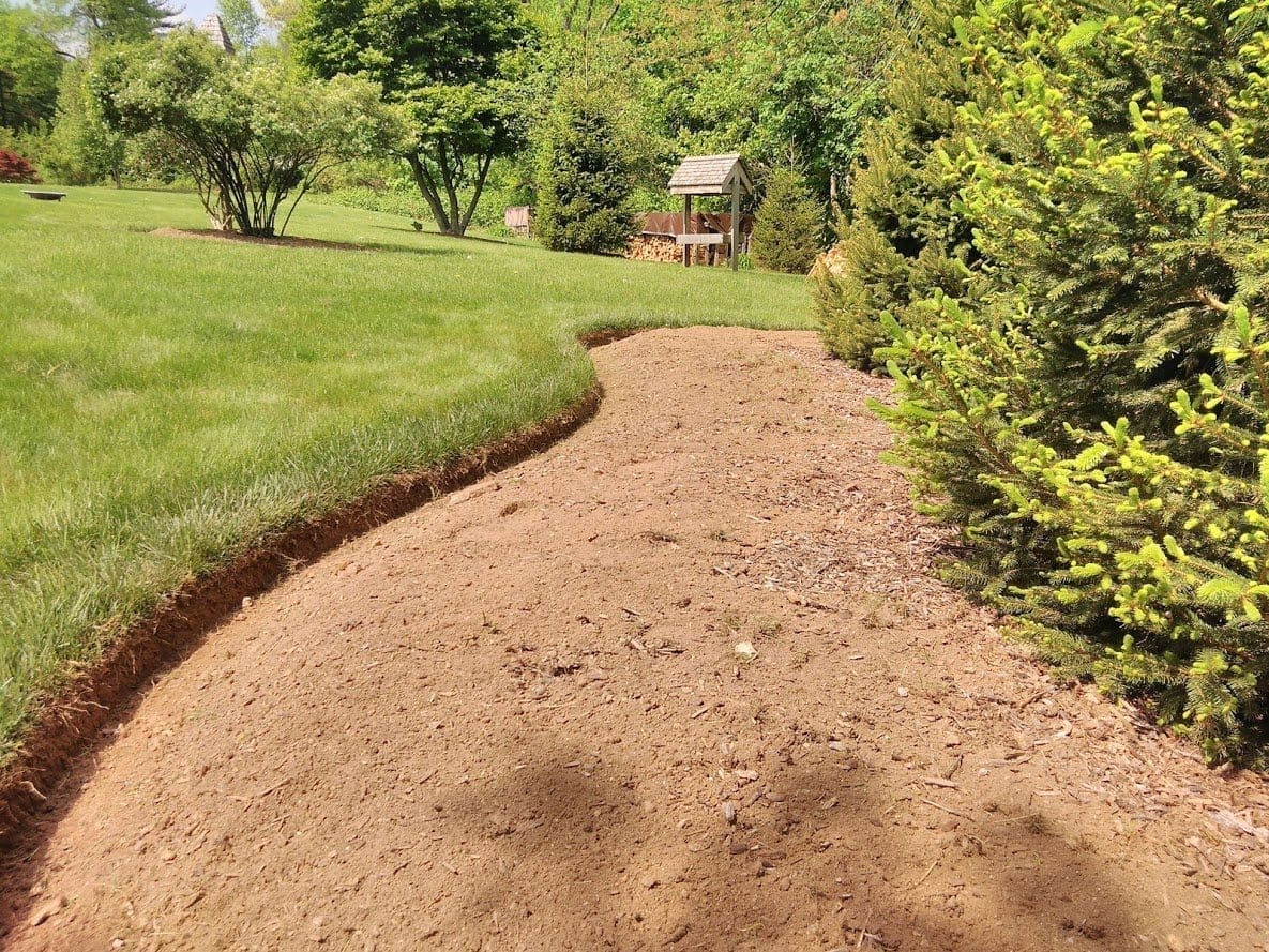 Outdoor Splendor - A freshly prepared garden bed with bare soil curves through a grassy lawn, bordered by green bushes and trees. A small wooden structure stands in the background amid lush greenery.