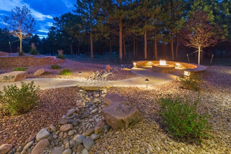 Outdoor Splendor - A landscaped backyard at dusk features a stone path with rocks, illuminated pathway lights, and a circular fire pit with built-in seating surrounded by trees.