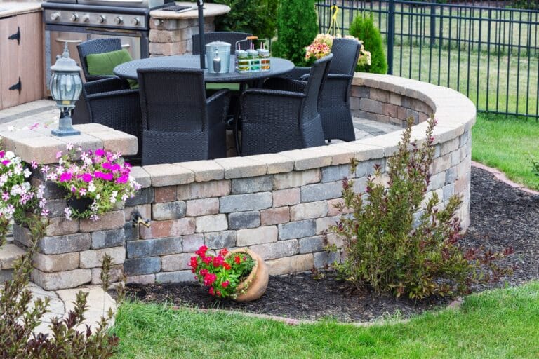 Outdoor Splendor - A backyard patio with a circular stone wall, black wicker chairs around a dark table, a grill, potted flowers, and landscaped garden beds with shrubs and grass.