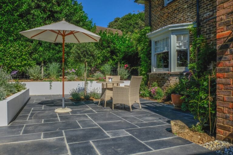 Outdoor Splendor - A sunny patio with dark stone tiles features a round table, two wicker chairs, and a large white umbrella. The area is surrounded by lush greenery, white planters, and part of a brick house with a bay window.