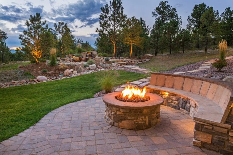 Outdoor Splendor - A curved stone bench surrounds a fire pit on a paved patio, set in a landscaped yard with green grass, trees, and a stone path at dusk.