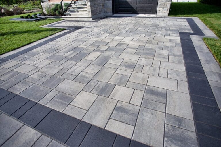Outdoor Splendor - Modern driveway with rectangular gray and dark gray interlocking pavers arranged in a geometric pattern, bordered by green grass and leading to stone steps and a garage.