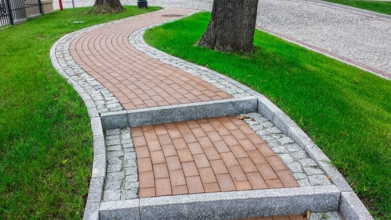Outdoor Splendor - A winding brick pathway with stone borders curves through green grass, featuring two small steps built into the path as it ascends beside large tree trunks.