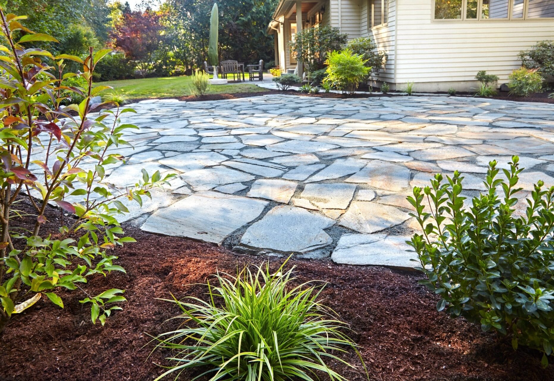 Outdoor Splendor - A backyard with a large flagstone patio surrounded by mulch and green plants, next to a beige house with a garden and lawn in the background.