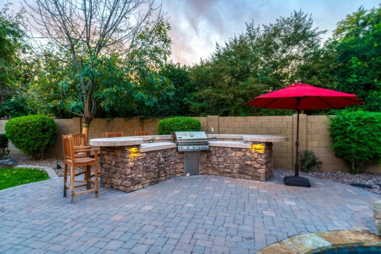 Outdoor Splendor - Outdoor patio with stone barbecue grill, two wooden bar stools, and a red umbrella. The area is surrounded by green trees, bushes, and a brick privacy wall, with paving stones covering the ground.