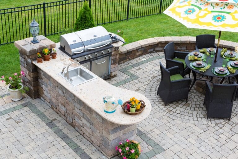 Outdoor Splendor - Outdoor patio featuring a built-in stone kitchen with a grill and sink, potted plants, and a nearby round dining table with place settings and an umbrella, all set on a patterned stone floor.