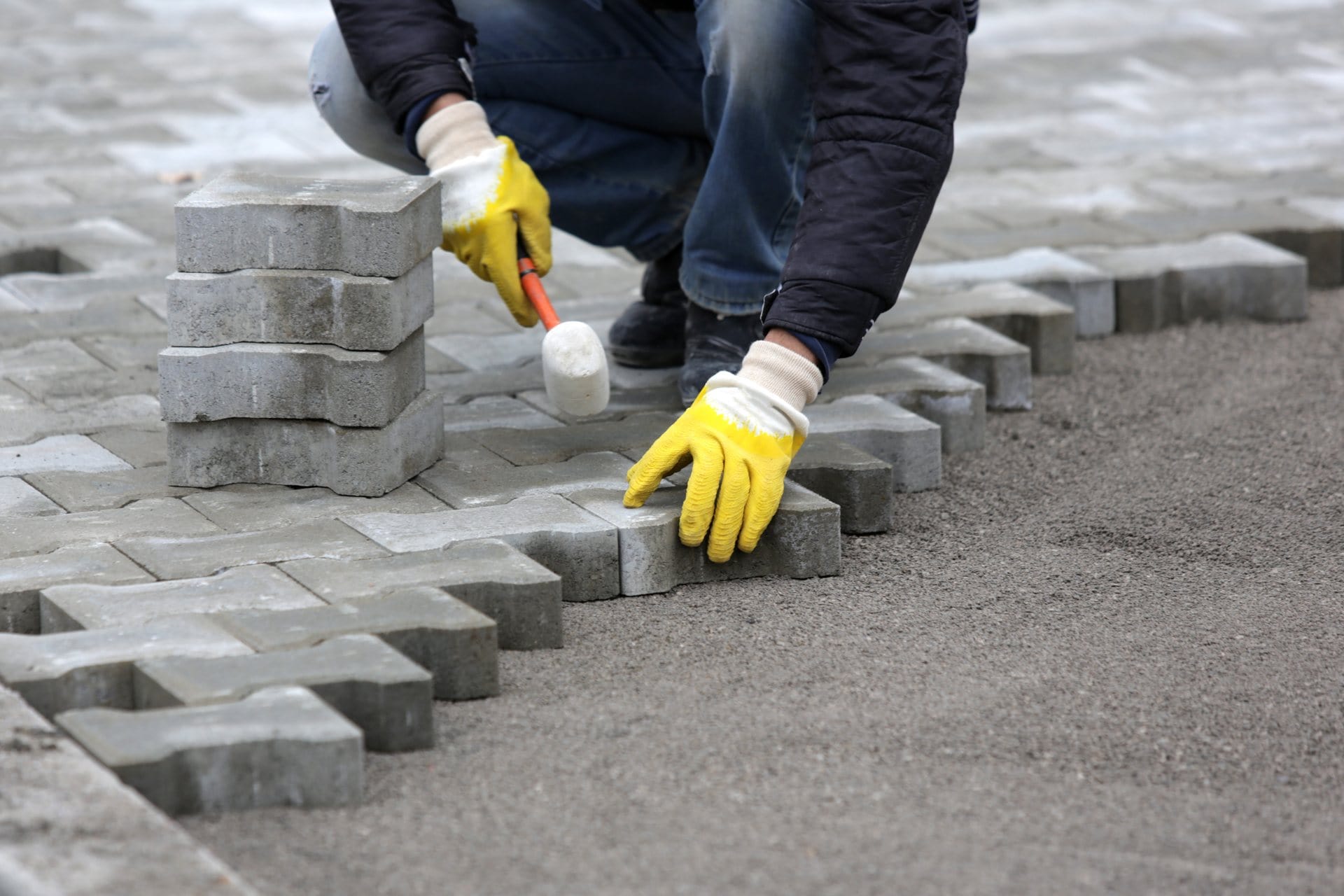 Outdoor Splendor - A person wearing yellow gloves uses a rubber mallet to lay gray paving stones in a pattern on the ground, working on a pavement or walkway construction project.
