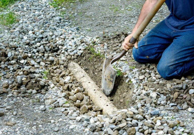 Outdoor Splendor - A person kneels on gravel and uses a shovel to dig around a partially buried pipe or log in the ground, surrounded by small and medium-sized rocks.