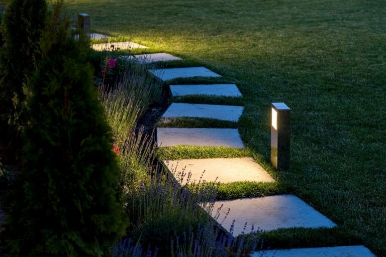 Outdoor Splendor - A stone pathway with rectangular stepping stones is lit by small, modern garden lights at dusk, bordered by green grass and shrubs.