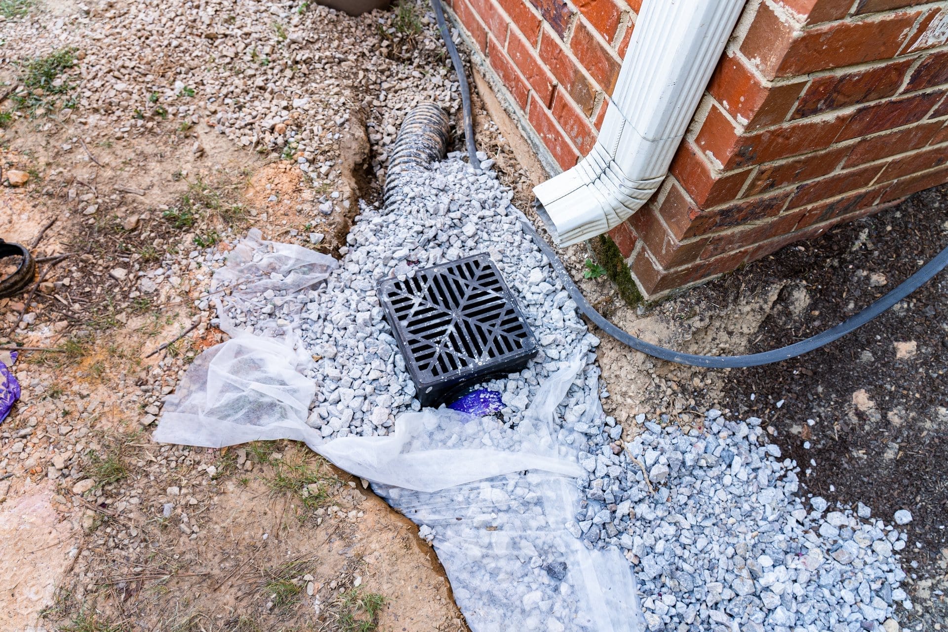 Outdoor Splendor - A black drainage grate sits on gravel next to a house with brick siding and a white downspout. Landscape drainage fabric and rocks surround the area, providing effective water management for the home.