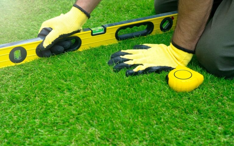 Outdoor Splendor - Person wearing yellow work gloves kneeling on grass, using a yellow spirit level and a yellow tape measure, checking the ground level.