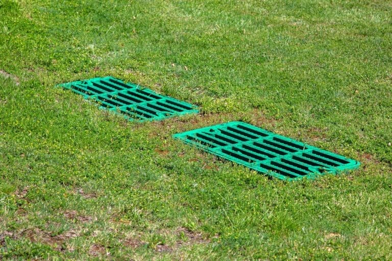 Outdoor Splendor - Two green storm drain grates are set into the ground amid green grass, allowing water drainage in an outdoor grassy area.