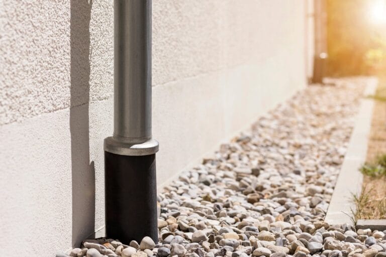 Outdoor Splendor - A metal downspout attached to the exterior wall of a building extends down into a black drain pipe, surrounded by light-colored gravel and stones, with sunlight illuminating part of the scene.