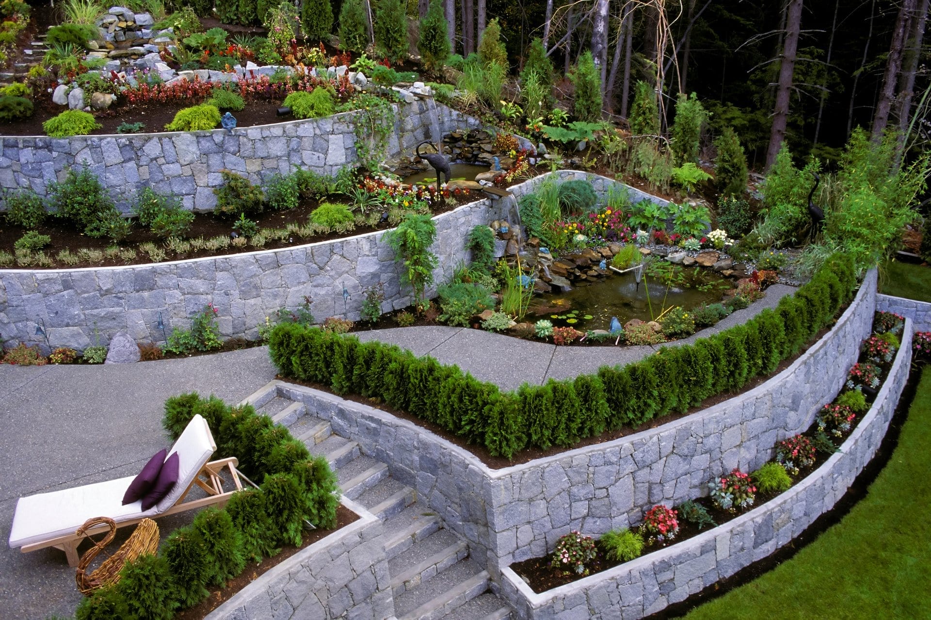 Outdoor Splendor - Tiered stone garden with lush green shrubs, flowering plants, and a small pond with a fountain. Curved stone walls separate the levels. A lounge chair with cushions sits on the patio near steps leading to the garden.