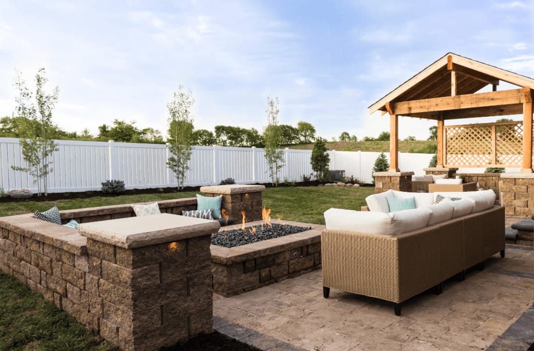 Outdoor Splendor - Transform your backyard with a modern patio featuring a built-in fire pit, cushioned seating, covered pergola, stone walls, and a white privacy fence—surrounded by green grass and young trees under a blue sky.