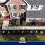 Outdoor Splendor - Aerial view of a brick house with a gray roof, landscaped front yard, mulched garden beds, and bushes showing recent spring pruning. A concrete walkway leads to the front door, with an "Outdoor Splendor" banner at the bottom.