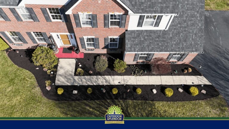 Outdoor Splendor - Aerial view of a brick house with a gray roof, landscaped front yard, mulched garden beds, and bushes showing recent spring pruning. A concrete walkway leads to the front door, with an "Outdoor Splendor" banner at the bottom.