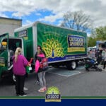 Outdoor Splendor - People gather around a green Outdoor Splendor landscaping truck at an outdoor event. Nearby, children and adults interact, and other vehicles are visible in the background on a cloudy day.