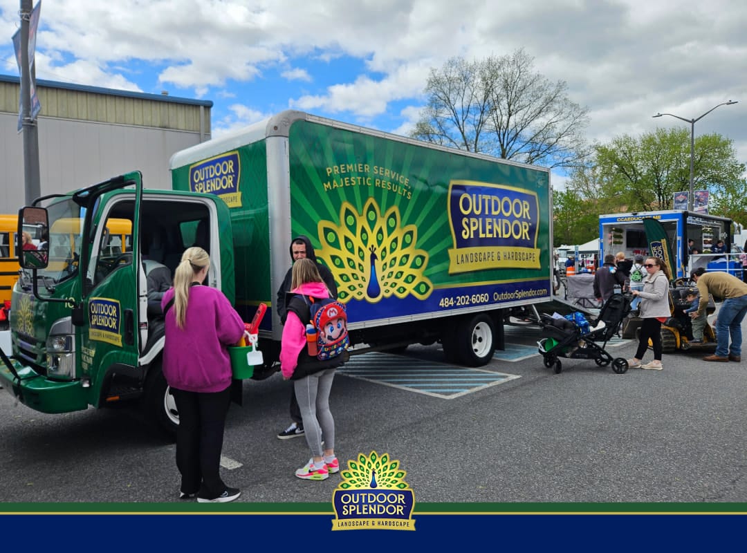 Outdoor Splendor - People gather around a green Outdoor Splendor landscaping truck at an outdoor event. Nearby, children and adults interact, and other vehicles are visible in the background on a cloudy day.