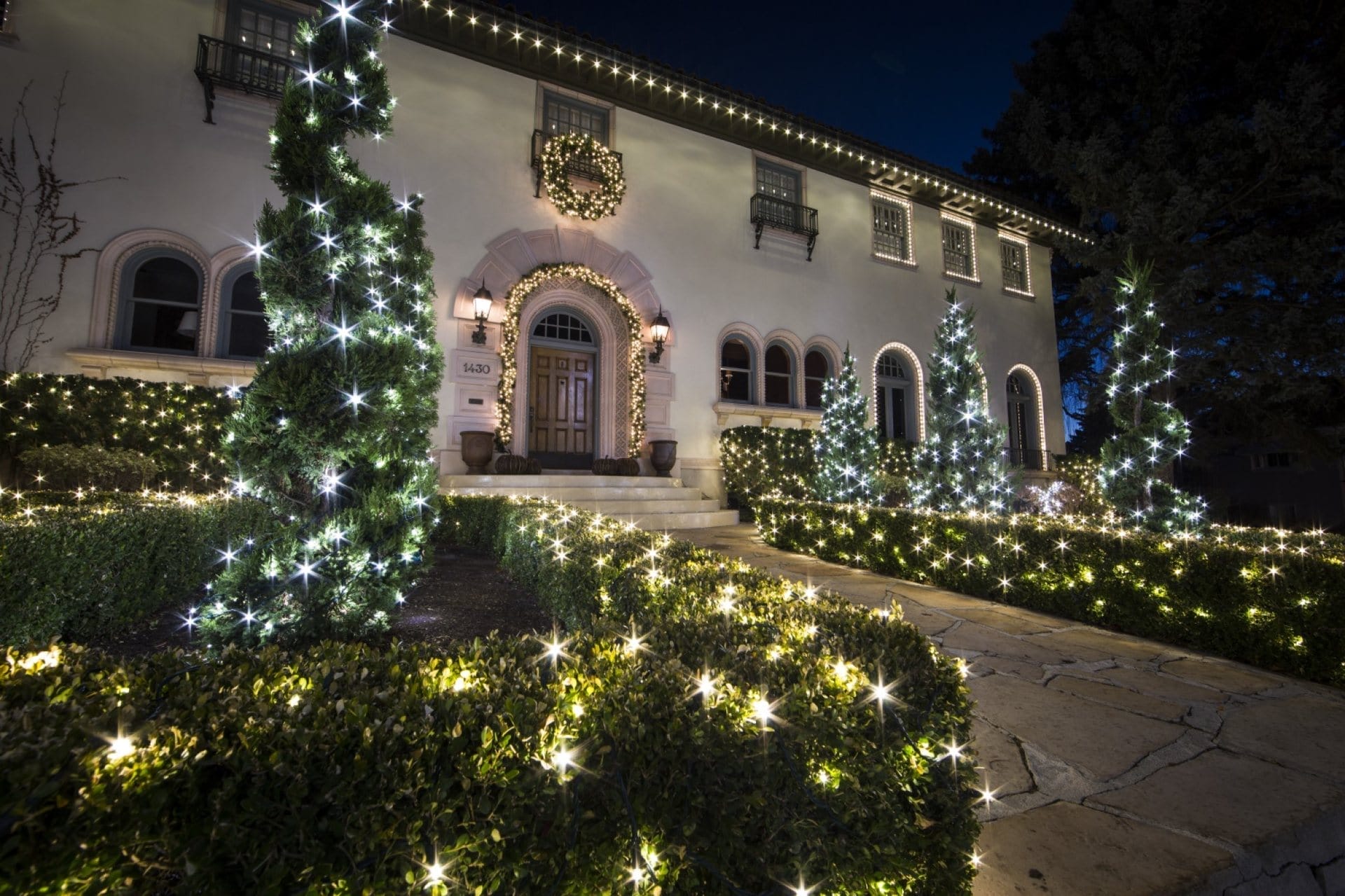 Outdoor Splendor - A large house decorated with white holiday lights along the roofline, bushes, and trees, with a lit wreath above the front door, creating a festive nighttime scene.
