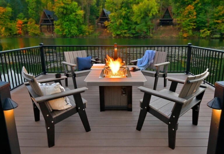 Outdoor Splendor - A cozy lakeside deck features outdoor furniture—four wooden chairs around a fire pit table, overlooking calm water with trees and cabins in the background. Blankets and pillows add comfort to the inviting seating area.